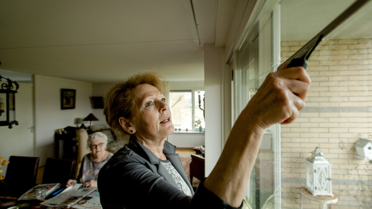 Een huishoudelijke hulp haalt een wisser over het raam, op de achtergrond zie je de bewoner van het huis een oudere dame.