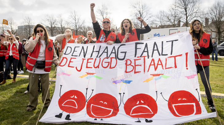 jeugdzorgmedewerkers voeren actie op het Malieveld, ze dragen oranje hesjes en houden een spandoek vast.