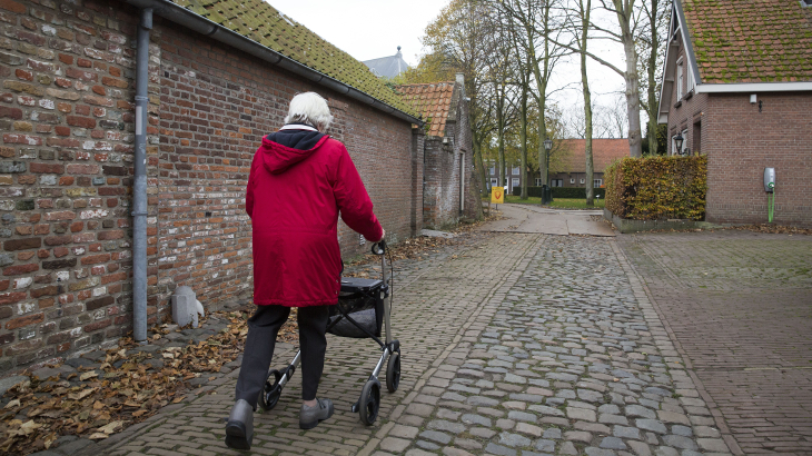 Een vrouw loopt met een rollator door een straat met oude bakstenen huizen en bomen.