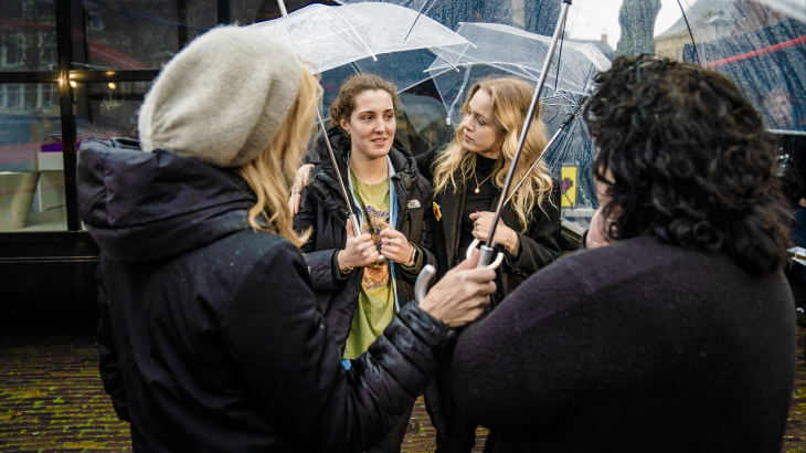 Twee jongeren met een doorzichtige paraplu praten met twee personen met een doorzichtige paraplu van wie je alleen de achterkant van het lichaam ziet.