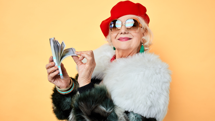 Een oudere vrouw in een bontjas, met een rode baret en met een grote zonnebril op, heeft een stapeltje bankbiljetten in haar handen.