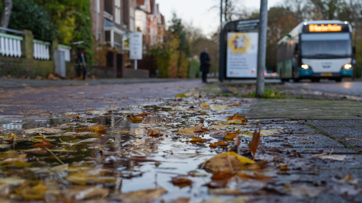 Een plas water op het fietspad met op de achtergrond een bushalte met een bus die arriveert.