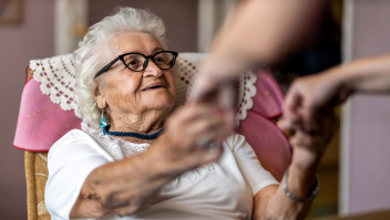 Een lachende oudere vrouw zit in een stoel en strekt haar handen uit naar iemand die niet in beeld is, op de handen na.