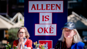 Twee zittende vrouwen met in het midden een affiche waarop staat: Alleen is oké.