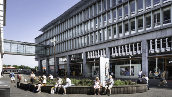 Mensen zitten in de zon op een muur voor het Maastrichtse stadskantoor in het winkel- en kantorencomplex Mosae Forum in het centrum van Maastricht.