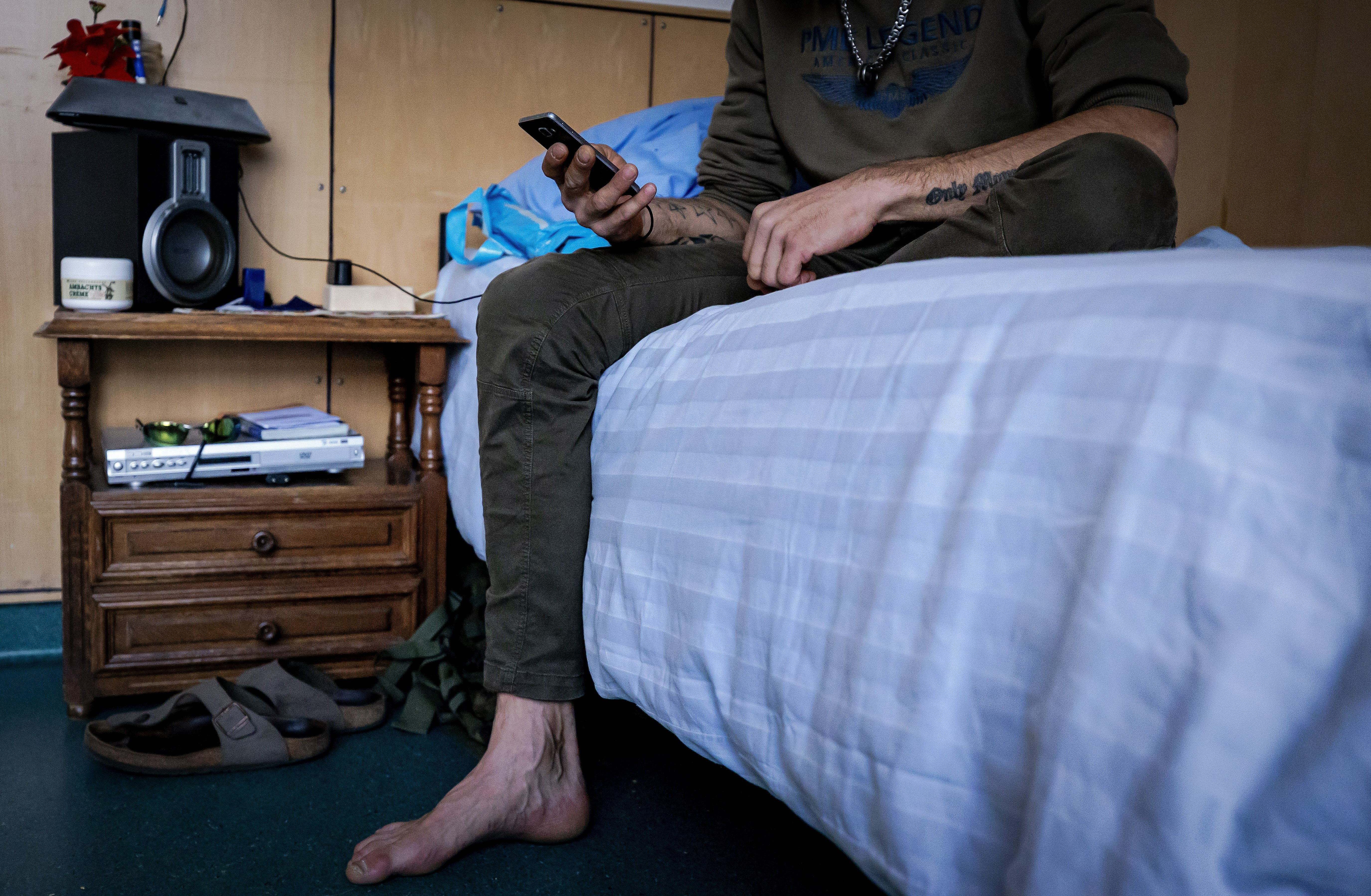 Een man met tatoeages zit op een bed en kijkt op zijn telefoon, je ziet zijn gezicht niet.